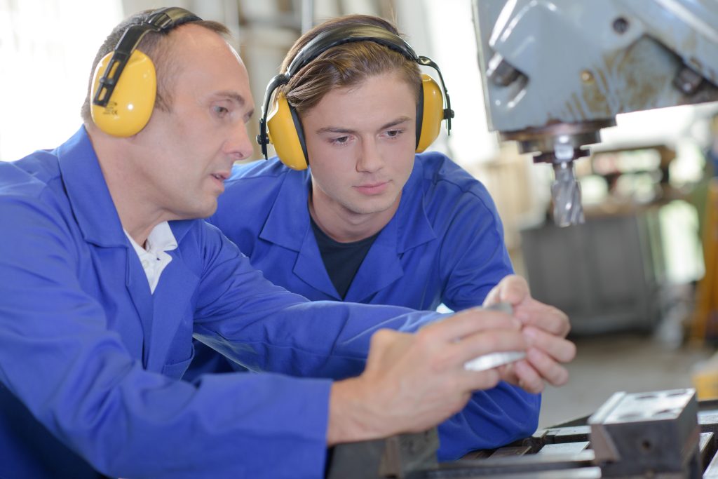 two males, apprentice and instructor, wearing ear protection working at machine